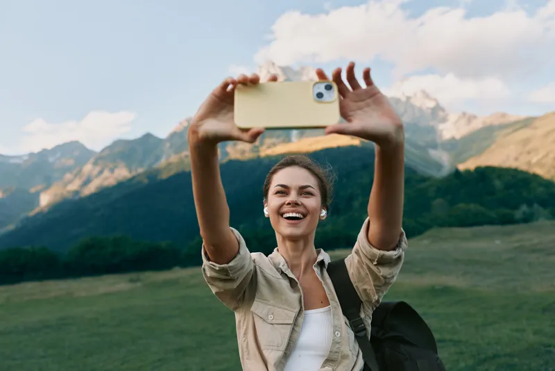 Young woman lifestyle traveler takes selfie with smartphone on mountain meadow background, smiling happy tourist enjoys nature and outdoor adventure in sunny day.