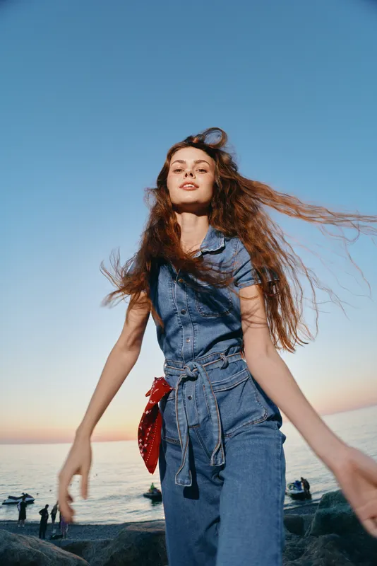 Lifestyle woman in modern boho-western denim street style with flowing hair captured in warm film color near the ocean during sunset, radiating free-spirited confidence.
