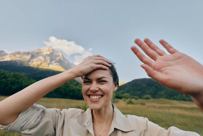 Joyful woman outdoors, warm sunlight, natural smile, hands in hair, relaxed pose, scenic mountains and green fields, bright mood, candid lifestyle moment