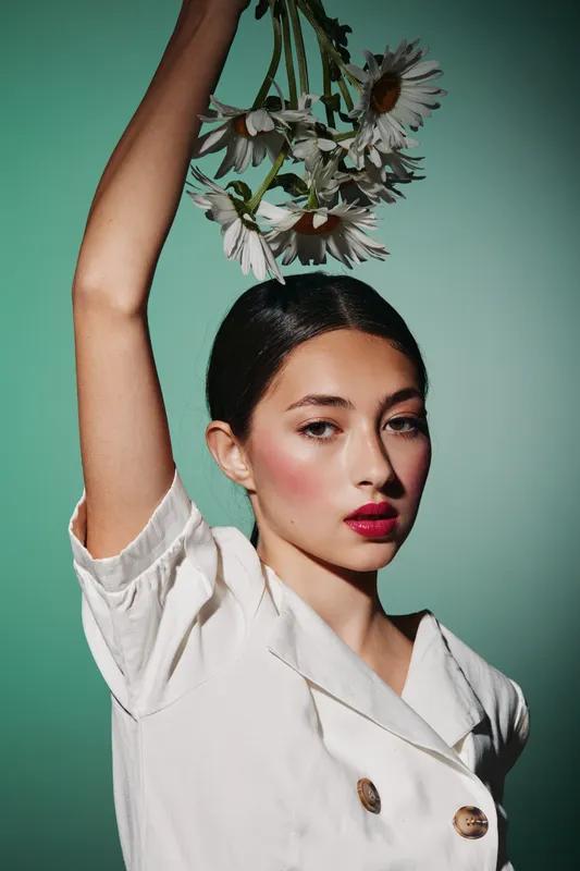 Young woman with vibrant makeup and red lips poses creatively holding daisies above head against green background. Fashion and beauty concept with artistic expression.
