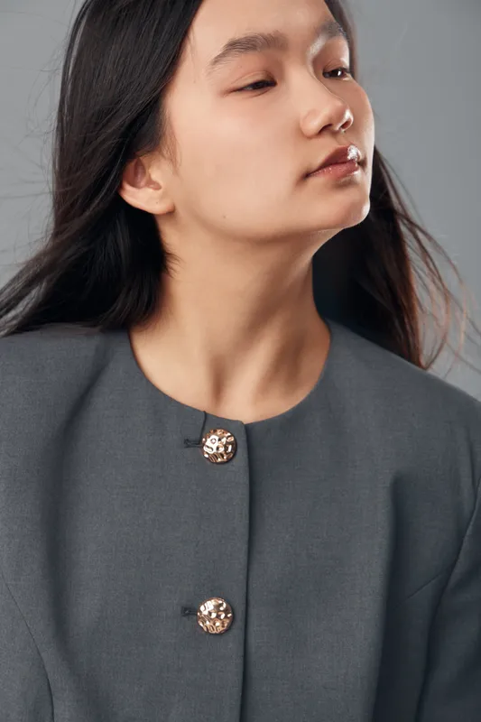 Young woman with long hair and closed eyes wearing gray buttoned jacket with decorative metallic buttons posing on neutral background for fashion portrait and style.