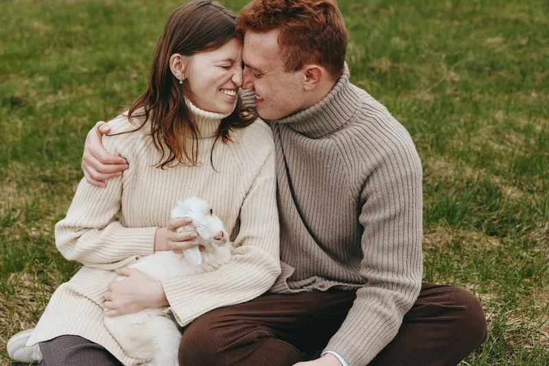 Young couple in cozy sweaters sits on grass holding a white baby goat while sharing a tender moment with smiles and closed eyes in a natural lifestyle setting.