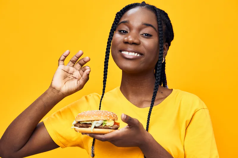 Happy young woman holding a delicious burger, smiling against a vibrant yellow background, showcasing fresh ingredients and joy for healthy eating