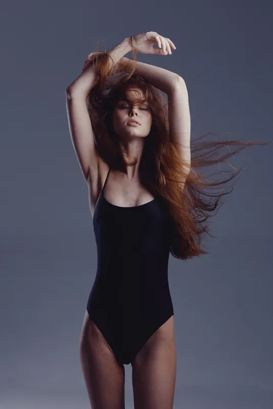 Young woman in black swimsuit posing with tousled hair against gray background, showcasing elegance and strength.