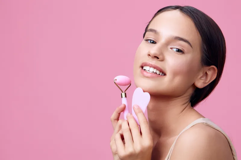 Young woman with a joyful expression holding a pink facial roller against a pink background, promoting self care and beauty