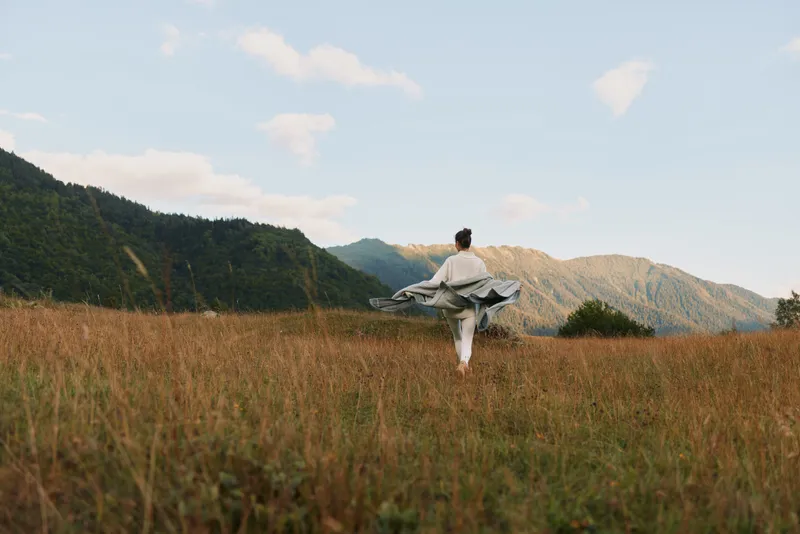 Young woman twirls a flowing dress in a sunlit meadow, mountains in the distance, embodying carefree outdoor style and natural movement as the breeze lifts the fabric.