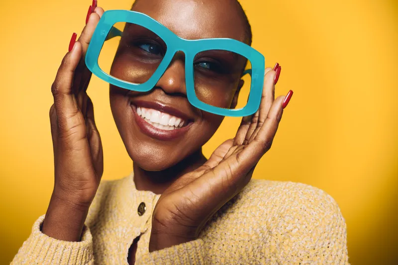 Happy dark skinned African woman with short hair wears oversized blue glasses and beige sweater smiling brightly on yellow background. Concept of joy, fashion, and beauty portrait.