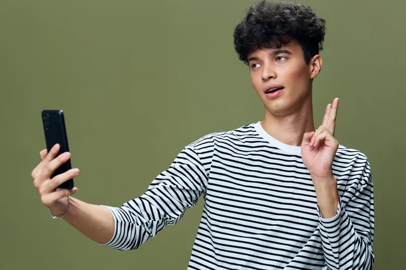 Young man taking selfie with smartphone while crossing fingers, wearing striped shirt against plain olive background, expressing hope and anticipation.