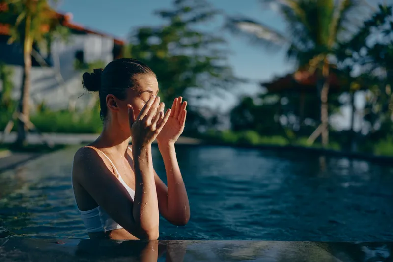 Woman in a sunlit pool, hands near face in a playful, relaxed moment, outdoor lifestyle scene with bright water, soft breeze, tropical backdrop, casual swimwear, candid leisure