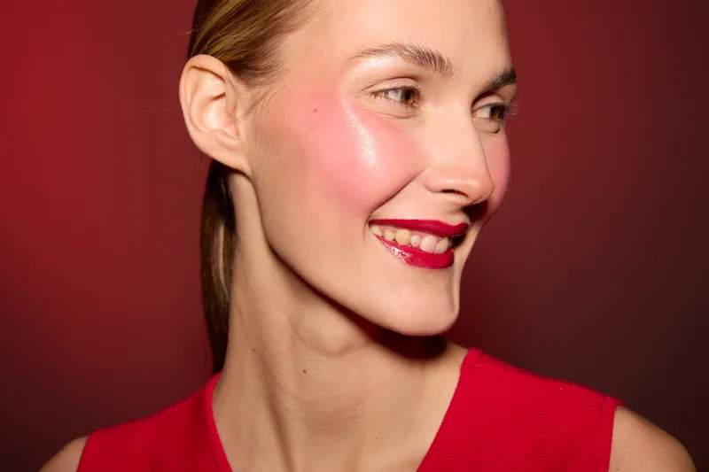 Smiling woman with shiny skin and red lipstick looks sideways on red background. Portrait of happy female model in red dress with natural makeup and glowing complexion. Beauty and fashion concept.