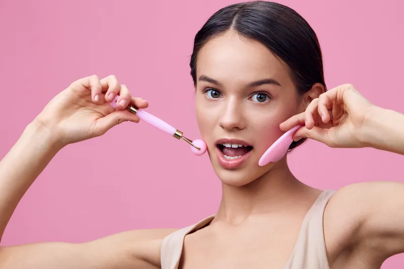 Young woman demonstrating facial roller for skincare on a pink background, expressing excitement and joy about beauty routine, perfect for wellness themes