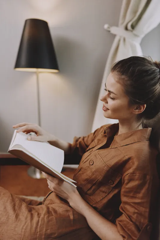 Молодая женщина в коричневом платье наслаждается чтением книги в помещении с мягким освещением, излучая спокойствие и тепло