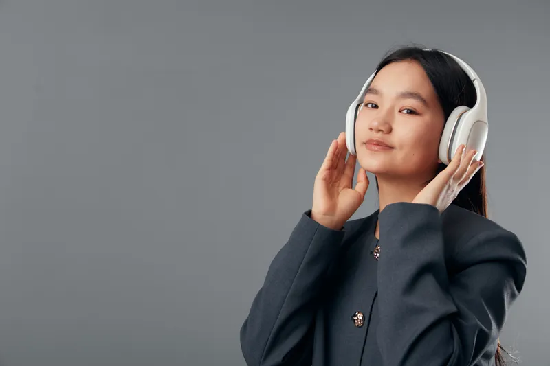 Young woman wearing wireless headphones and dark jacket listens to music with closed eyes on neutral gray background, enjoying sound and relaxation indoors with calm expression.