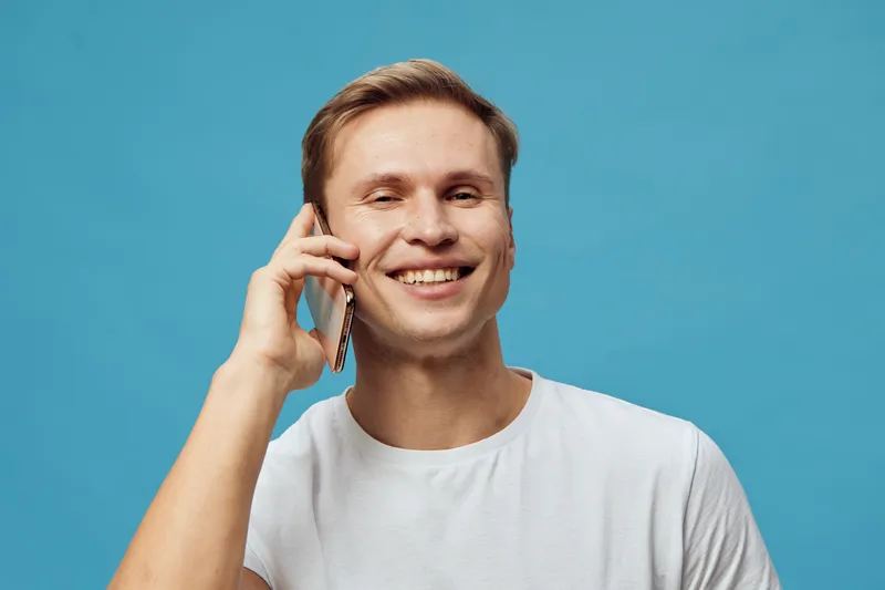 Smiling man talking on phone, looking happy and relaxed, casual white t-shirt, blue background, studio portrait, lifestyle concept, communication, happiness, technology, modern, leisure.