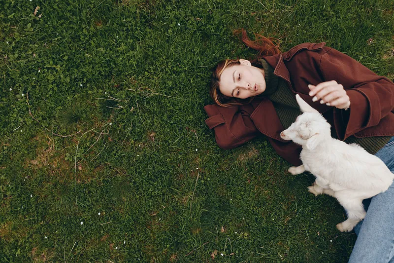 Relaxed woman lying on grass with a playful goat kid beside her, in a sunny park setting. Casual lifestyle scene showcasing companionship, wellness, and outdoor living. Soft light and natural colors