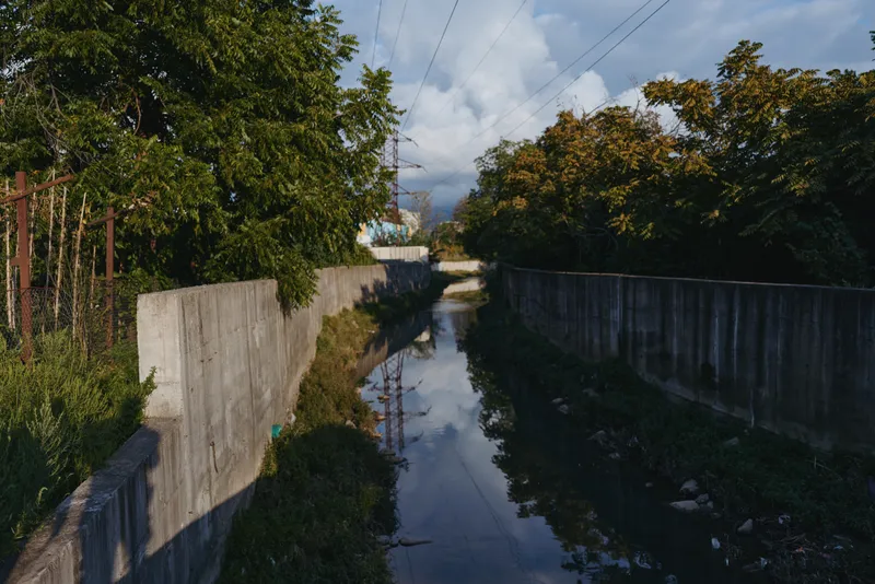 Urban canal with concrete channel and river reflection, flanked by overgrown vegetation and power lines under a cloudy sky in an industrial landscape, moody travel or environment stock.