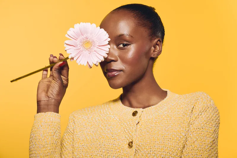 Dark skinned African woman holds pink flower close to her eye, wearing yellow sweater against yellow background. Concept of beauty, nature, self care, and floral aesthetics.