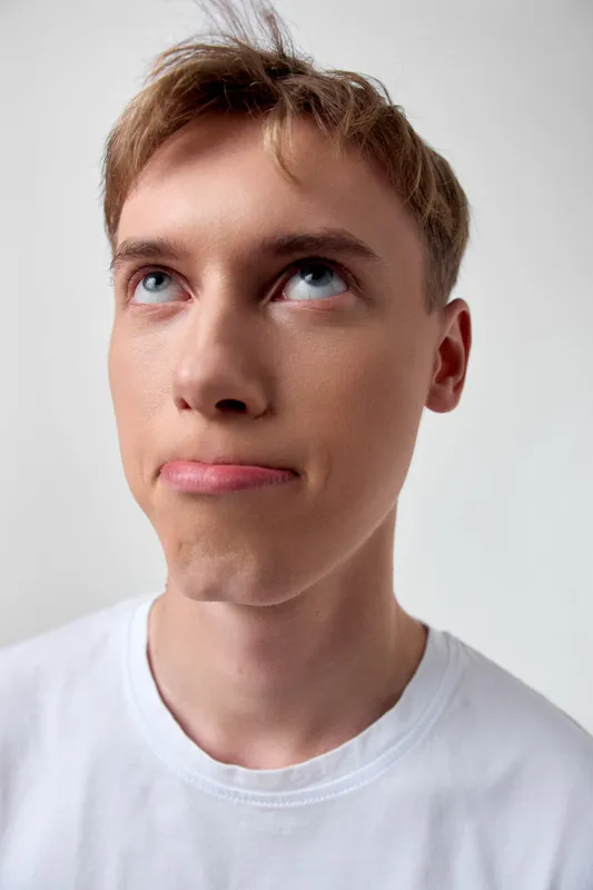 Young man with upward gaze and curious expression wearing a white t shirt in a studio shot with soft lighting and a minimal background, delivering thoughtful, modern stock imagery