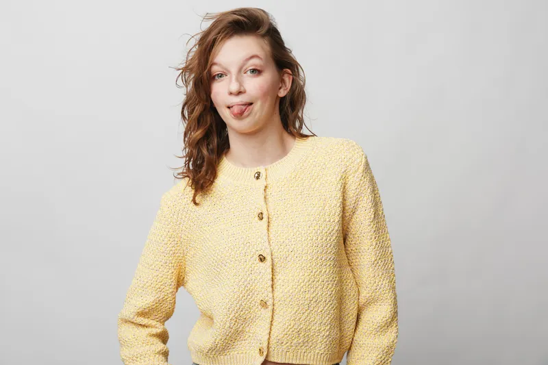 Young woman in yellow knitted cardigan makes funny face sticking out tongue. Casual style, playful mood, isolated on light grey background. Concept of humor, personality, and self-expression.