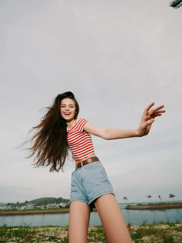 Playful young woman poses energetically by a seaside landscape, wearing a red and white striped tee and denim shorts, smiling with windblown hair under a bright sky.