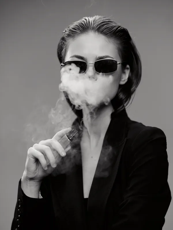Black and white portrait of a woman vaping with sunglasses and a dark jacket in studio lighting.