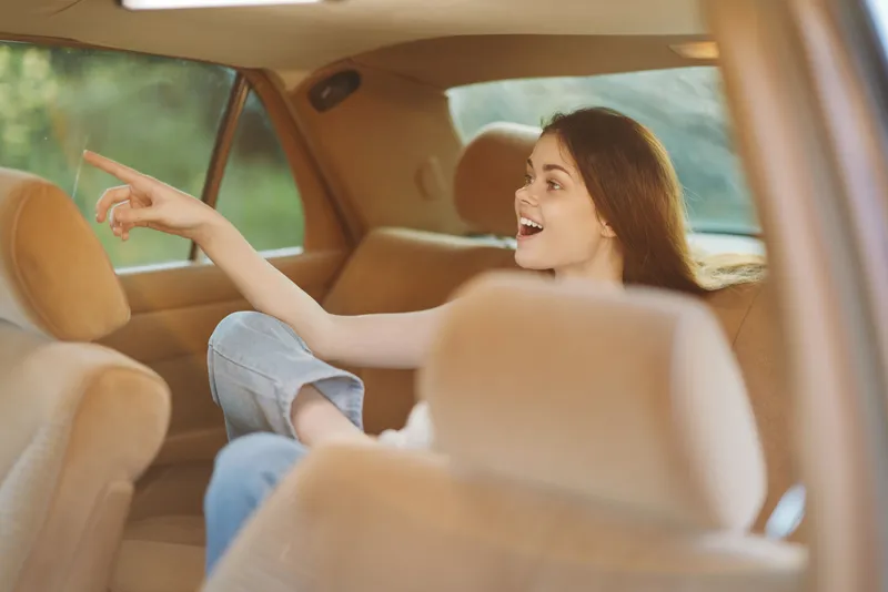 Young woman enjoying a road trip, pointing out the window with joy, wearing casual clothing, inside a car, with a vibrant green background.
