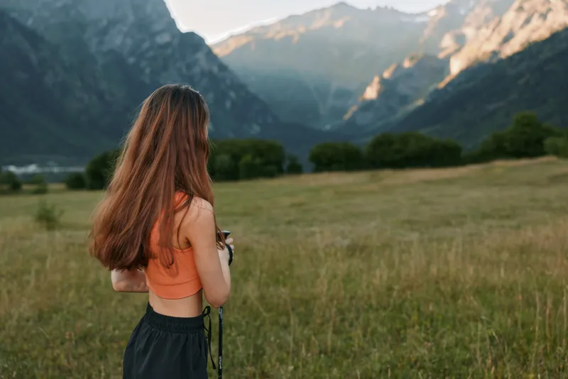 A lone photographer stands in a grassy field with a mountain range in the distance, capturing the landscape with calm focus and a sense of outdoor adventure.