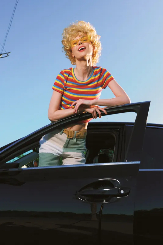 A cheerful woman leaning from a sleek car window, wearing a striped shirt and denim, under a bright sky. Retro, sunny lifestyle portrait with fashion, color, and casual vibe.