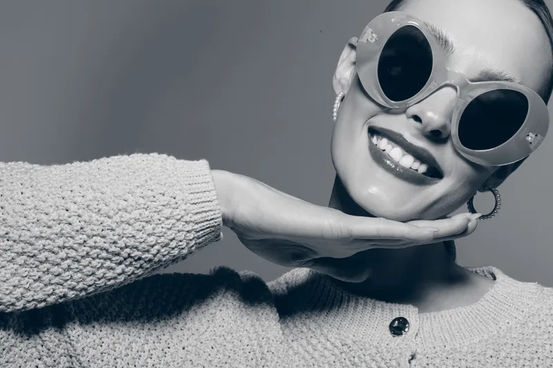 Smiling African American woman with dark skin wearing large round sunglasses and knitted sweater poses with hand under chin, playful mood.