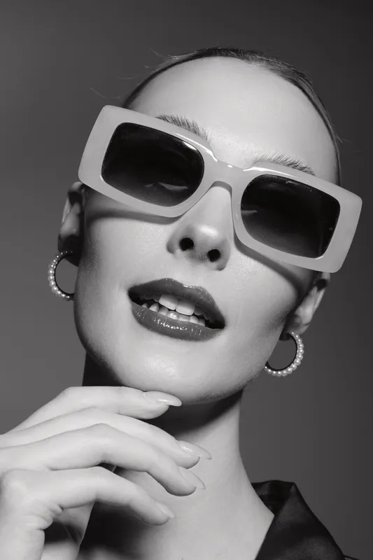 Close-up portrait of a stylish white woman with pale skin wearing large sunglasses and hoop earrings. Fashion concept with confident expression in black and white studio lighting.