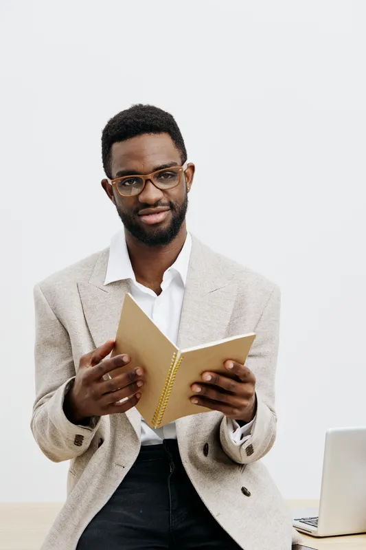 Confident young man with glasses standing against plain white background, holding open notebook, dressed in smart casual beige blazer, looking directly at camera, indoor studio portrait. People lifest