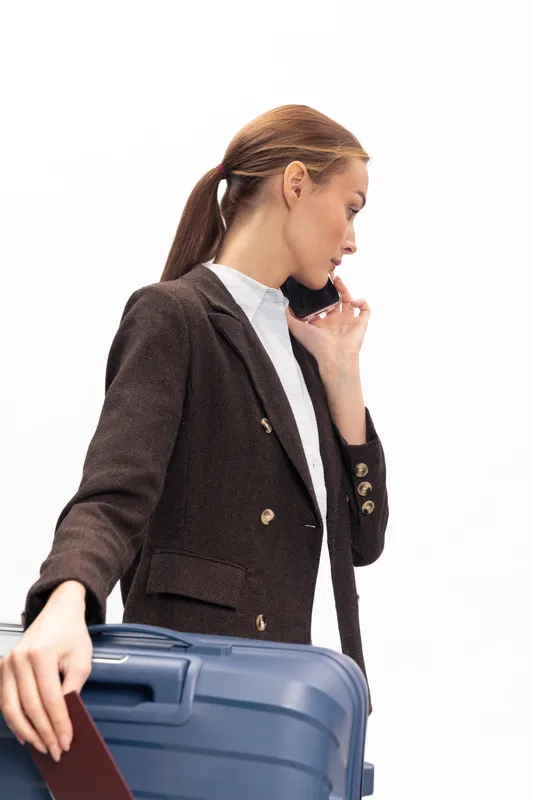 Young woman in brown blazer holds blue suitcase and talks on smartphone. She stands isolated on white background with passport in hand. Concept of travel, business trip, and communication.