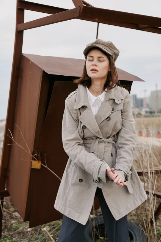 Young woman in beige trench coat and cap stands outdoors near rusty metal structure, eyes closed and hands clasped. Concept of autumn fashion, contemplation, and urban exploration.