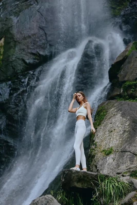 Woman standing near waterfall in white activewear, surrounded by rocks and lush greenery, enjoying serene nature and peaceful outdoor environment.
