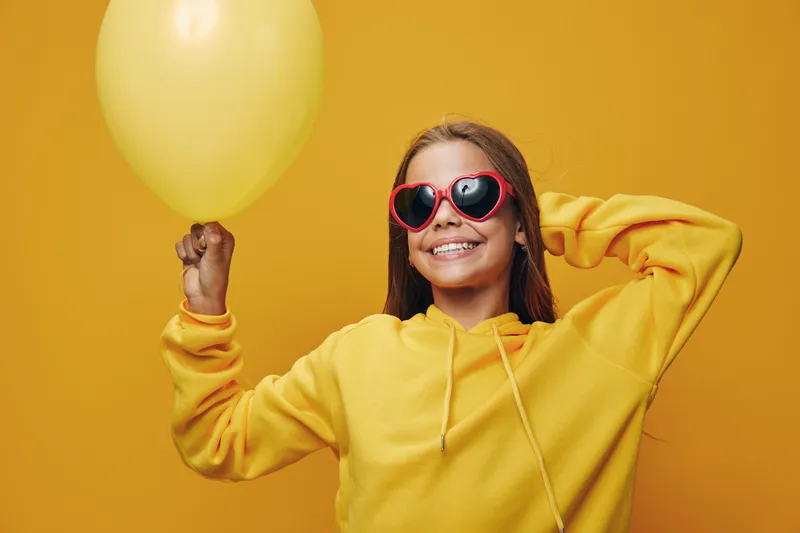 Girl smiling and holding yellow balloon in hand wearing heart-shaped sunglasses and yellow hoodie with bright background for happy mood and celebration.