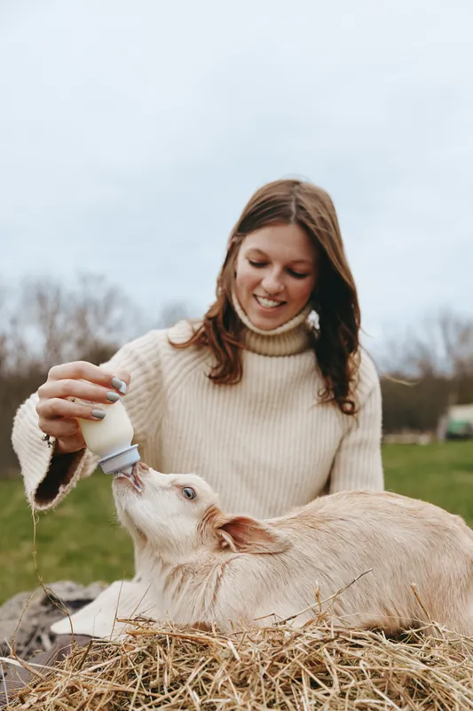 Young woman in a beige sweater feeds a baby goat with a bottle while sitting outdoors on a farm in a relaxed lifestyle scene.