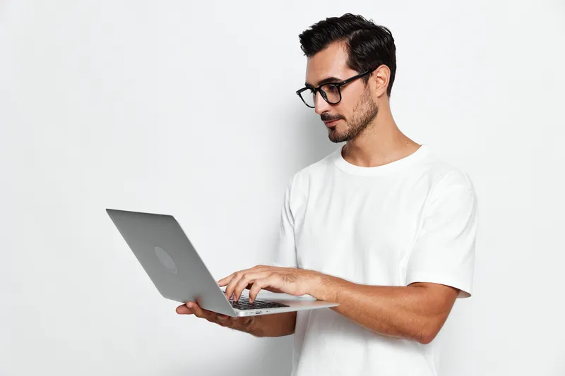 Stylish young man with glasses working on laptop, looking focused and determined while standing against a plain white background. Modern lifestyle concept.