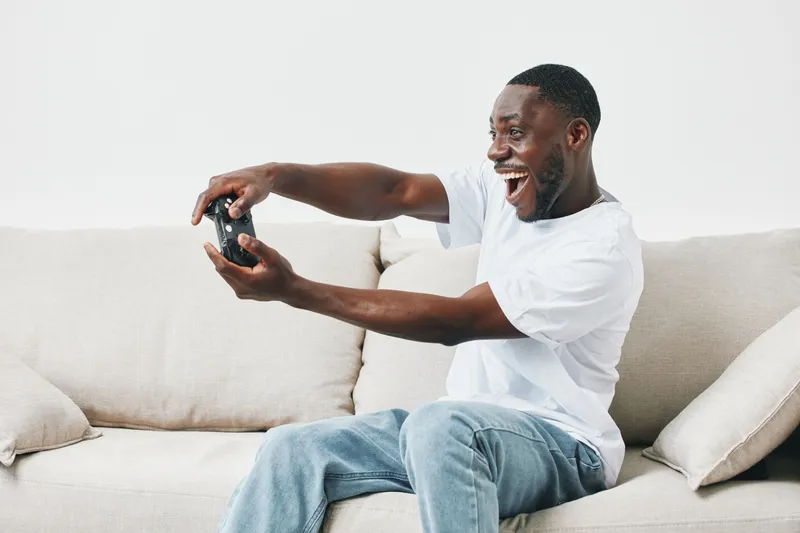 Joyful African man playing video games on the couch, expressing excitement with a game controller, wearing a casual white t shirt and jeans