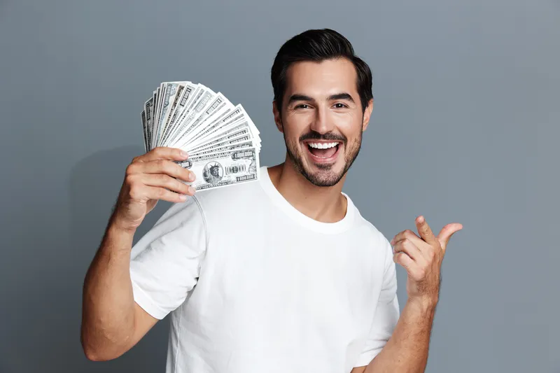 Happy smiling man holding fan of cash and pointing at camera with excitement. Confident joyful expression set against a grey background. People finance lifestyle concept.
