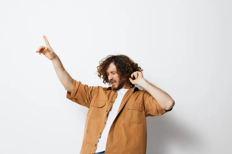 Young man with curly hair dancing joyfully in a stylish casual outfit against a bright, minimal background, expressing happiness and freedom
