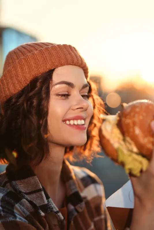 woman smile burger beanie sunset candid authenticity lifestyle. Young woman in warm beanie bites a burger at golden hour glow, candid lifestyle portrait with emotional storytelling and mindful living