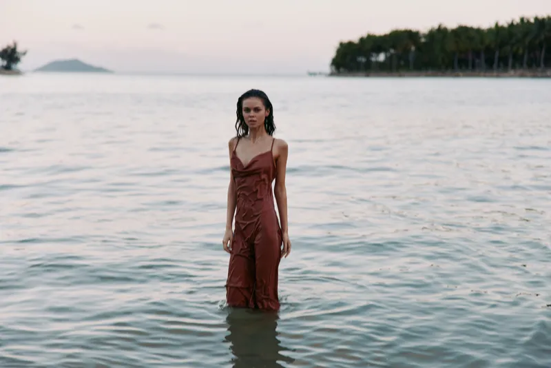 A solitary woman stands waist deep in calm sea water, wearing a rust-colored dress as soft light fades, creating a serene, contemplative beach scene.
