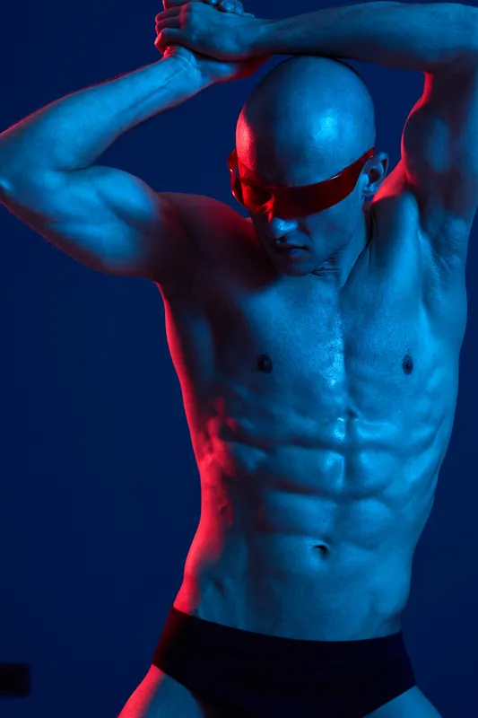 Athletic man with sunglasses striking a powerful pose, showcasing his muscular physique against a vibrant blue and red background in a captivating display of strength.
