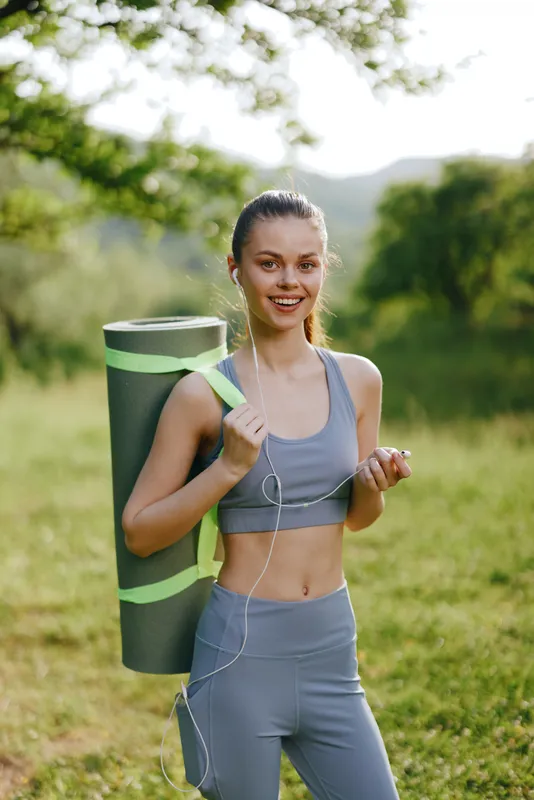 Fit young woman with yoga mat, smiling outdoors in sportswear, enjoying fitness exercise in nature, ideal for healthy lifestyle concepts