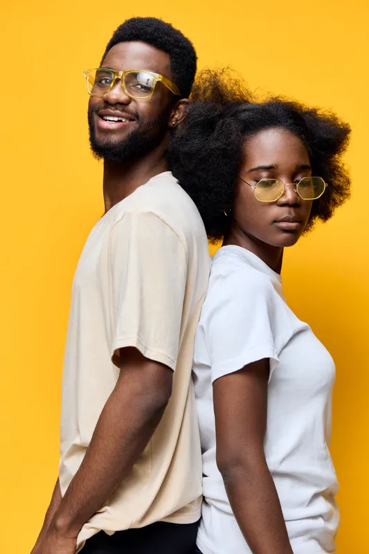 A stylish young couple posing back to back against a bright yellow background, showcasing trendy outfits and glasses, conveying confidence and happiness
