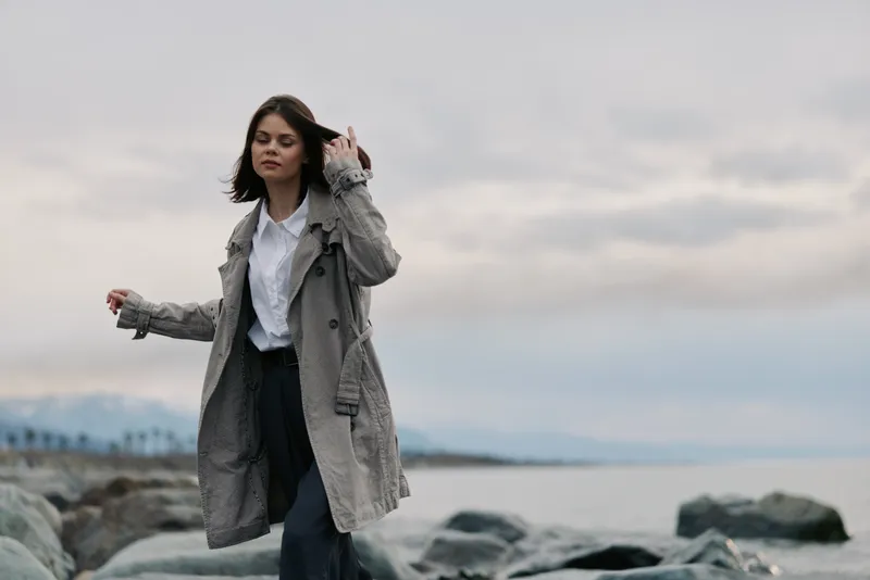Young woman in casual coat walking on rocky beach near calm sea under cloudy sky. Concept for nature, fashion, freedom, and peaceful outdoor lifestyle.