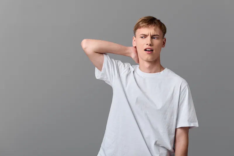 Young man in a white tee stretches his neck with a puzzled expression in a minimal studio, conveying fatigue, discomfort, and everyday hesitation
