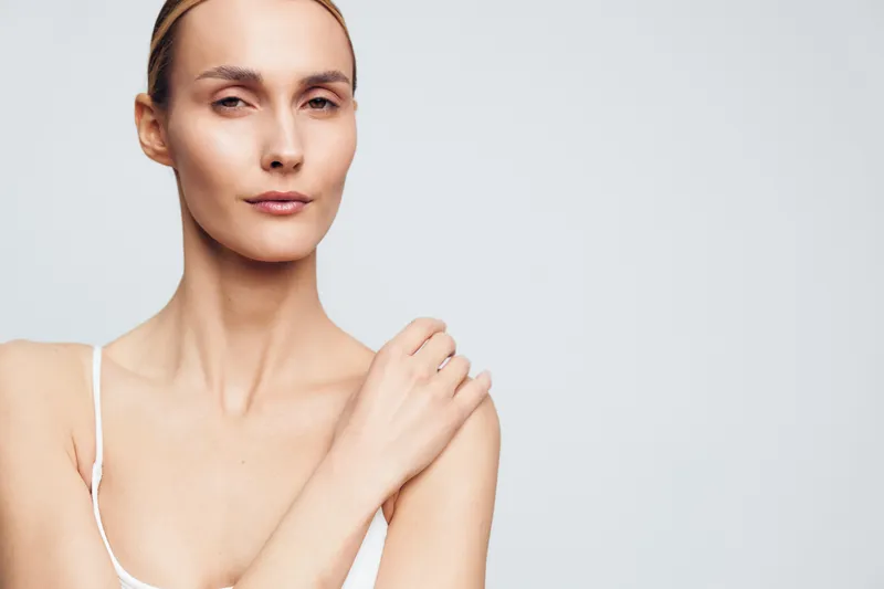 Portrait of confident woman with natural skin, minimal makeup, neutral expression, posing with hand on shoulder, soft lighting, plain background, beauty concept
