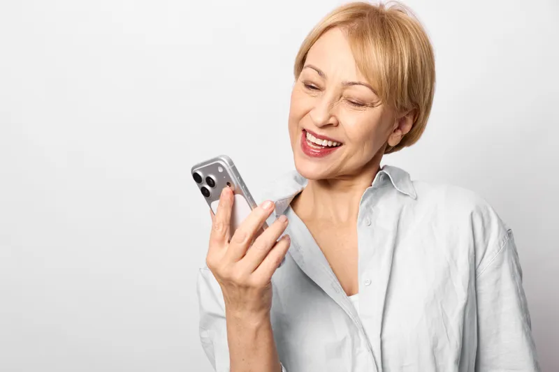 Smiling senior woman using smartphone in a studio portrait, indoors on a white background, joyful expression, communication and technology concept, soft light, candid moment