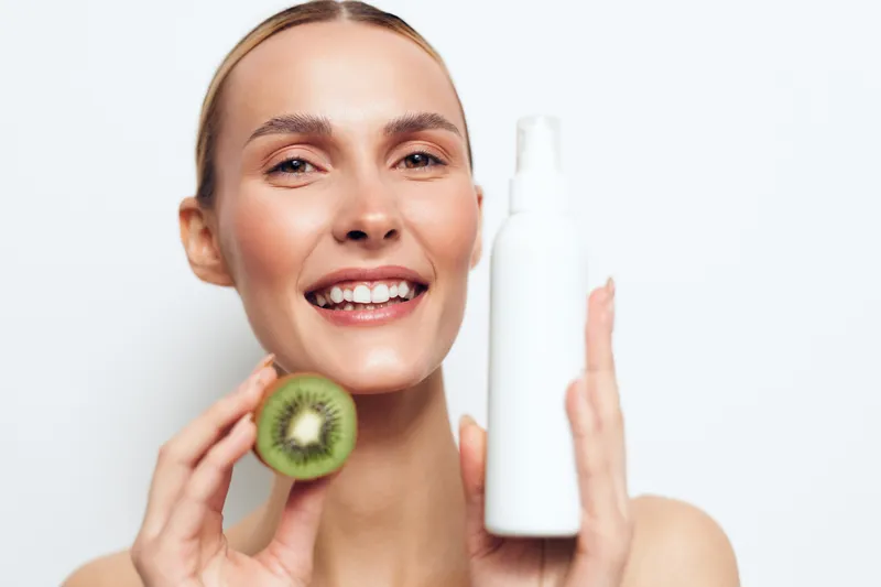 Woman, smiling, kiwi, skincare, face, cream, self, care, wellness, studio lighting, close up portrait, holding kiwi slice and cosmetic cream bottle, concept of hydration and natural beauty